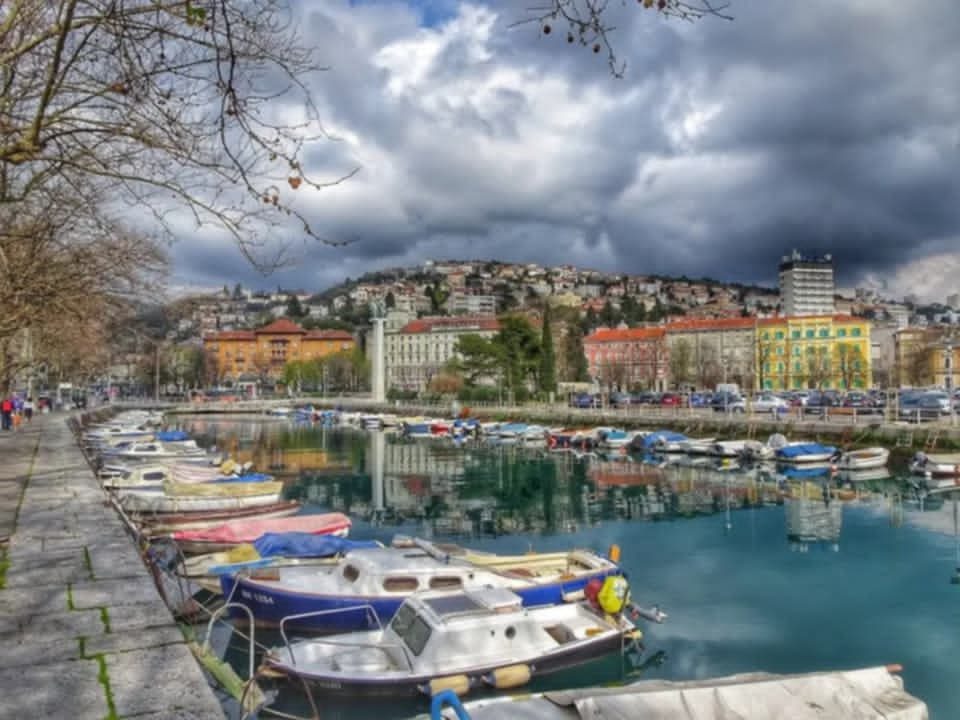 Brodice vezane u Mrtvom kanalu u Rijeci s pogledom na šarene zgrade i brda u pozadini pod oblačnim nebom.