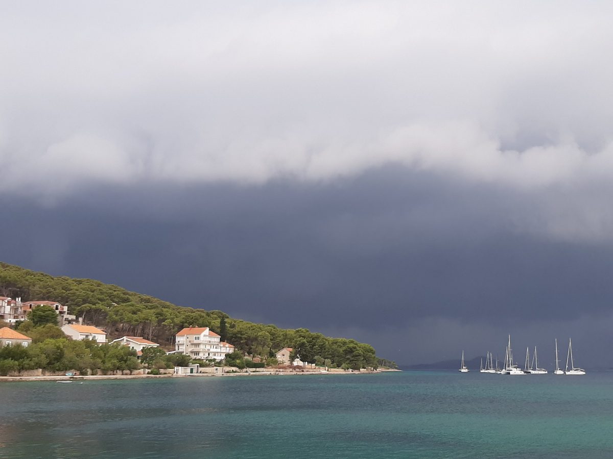 Pogled na obalu otoka Zlarina s kućama i zelenilom pod gustim, tamnim olujnim oblacima.